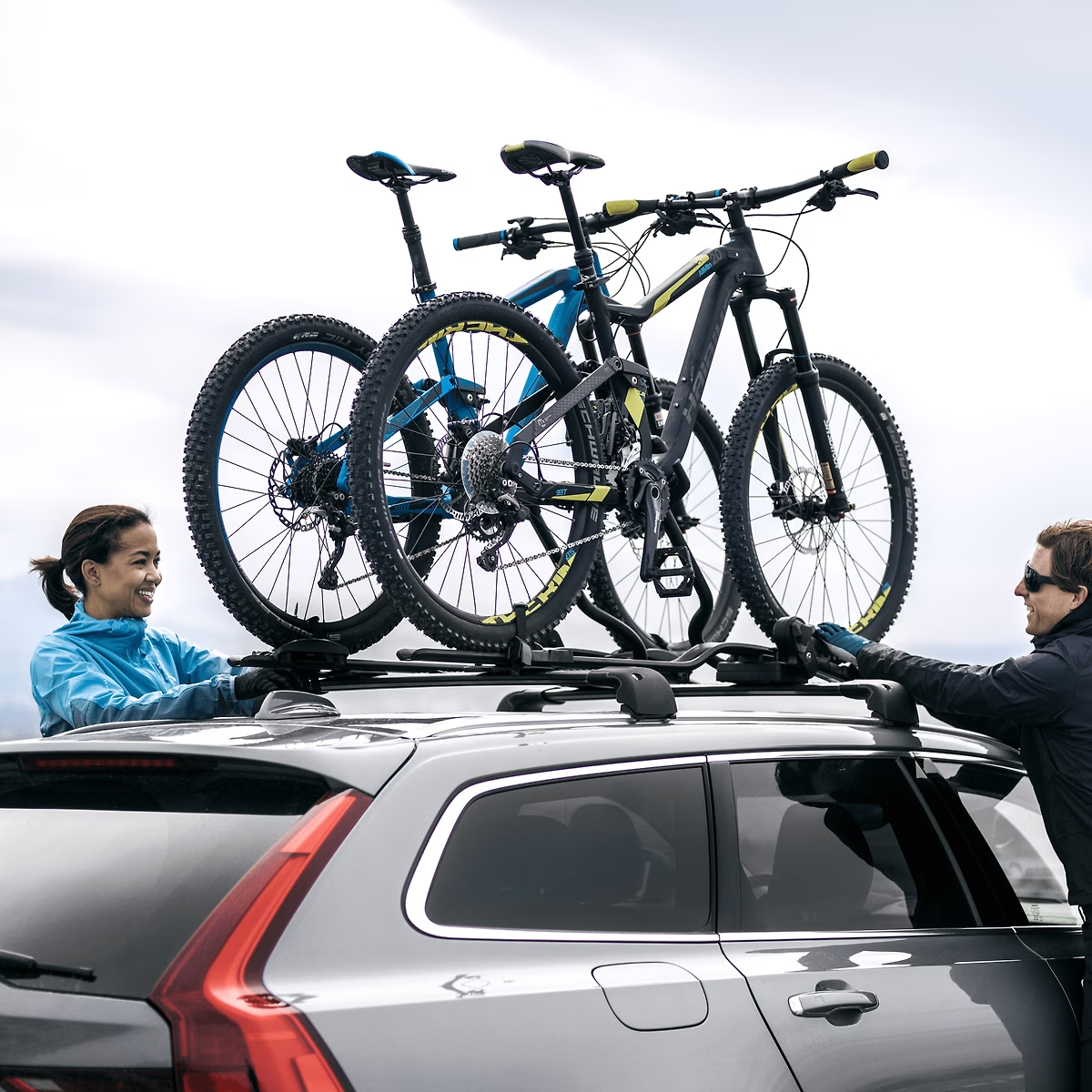 Two people loading bicycles onto a car roof rack against a cloudy sky.